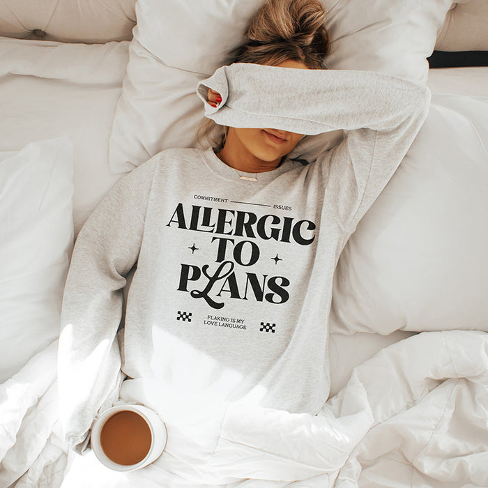 Woman lounging in bed wearing the Allergic to Plans ash grey sweatshirt, holding a coffee mug — cozy introvert aesthetic.