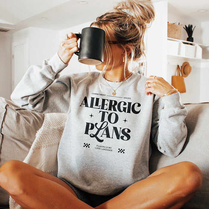 Woman with messy bun holding coffee while wearing the Allergic to Plans sweatshirt in athletic heather — cozy, stay-home vibes.