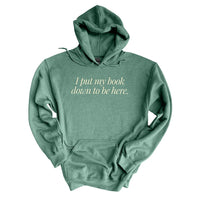 Flat lay of heather dark green “I Put My Book Down To Be Here” hoodie – funny reading hoodie for bookworms
