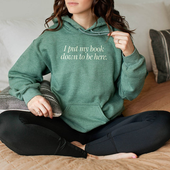 Woman wearing heather dark green “I Put My Book Down To Be Here” hoodie – funny bookish pullover for avid readers