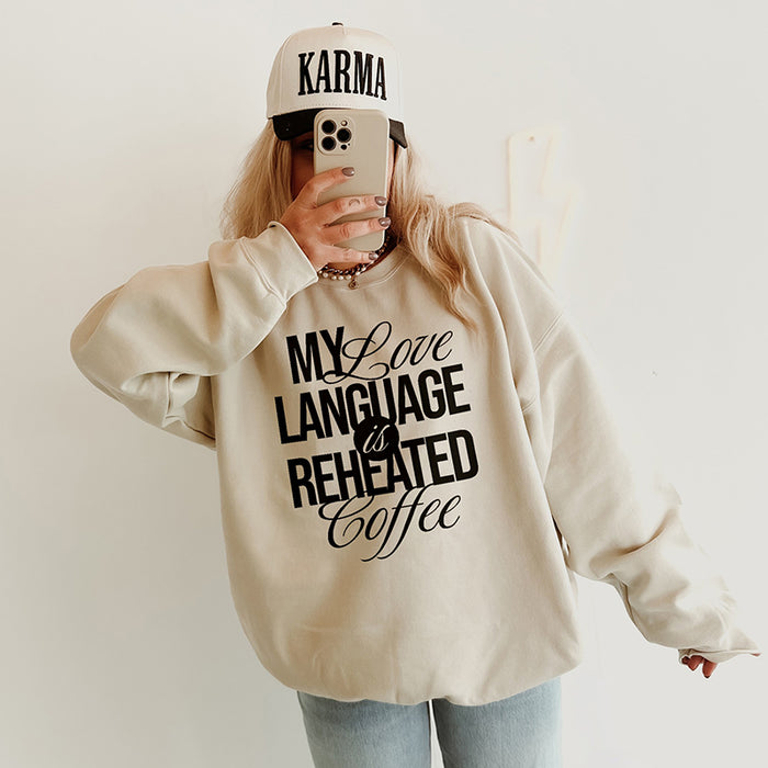 Woman wearing the My Love Language Is Reheated Coffee sweatshirt in sand (Gildan style) with jeans and a trucker hat — casual coffee lover outfit.