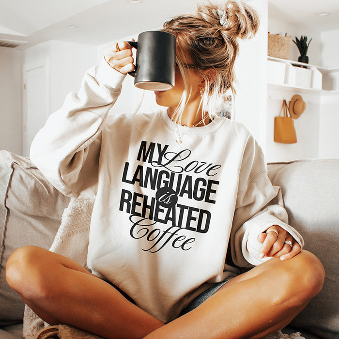 Model with messy bun sitting on a couch, wearing the My Love Language Is Reheated Coffee sweatshirt in sand and holding a coffee mug — cozy homebody vibes.