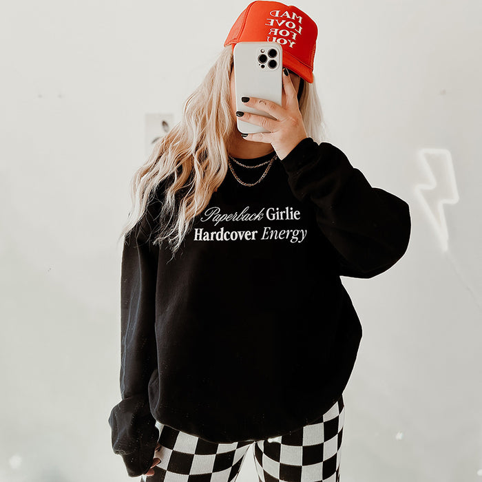 Model in black Paperback Girlie Hardcover Energy sweatshirt (Gildan style) styled with a trucker hat and black-and-white checkered pants — casual bookish vibes.