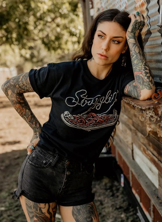 Woman with dark hair and tattoos wearing Strugglin’ graphic tee with black denim shorts. Casual outfit, skeleton and pasta graphic.