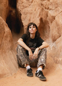 dark-haired model sitting in camo pants, wearing the Strugglin’ tee. Casual, outdoorsy vibe.