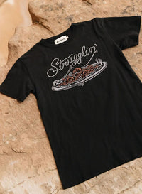 Flatlay of Strugglin’ graphic tee on outdoor background. Skeleton and spaghetti design centered on shirt.