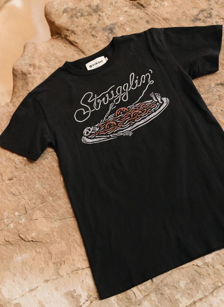 Flatlay of Strugglin’ graphic tee on outdoor background. Skeleton and spaghetti design centered on shirt.